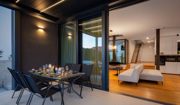 A dining set on the terrace with electronic pergola system and a view on the interior of the Villa with living room and kitchen