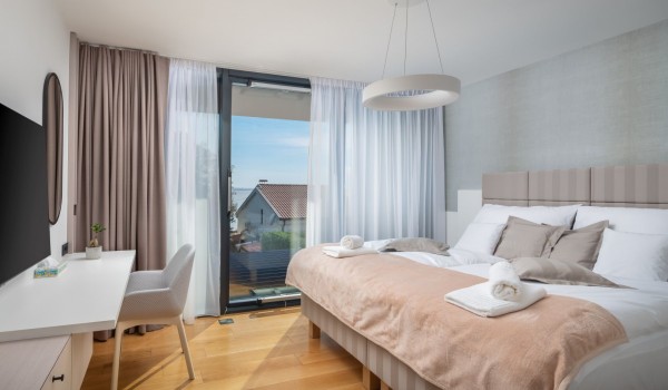 Modernly decorated bedroom with a king size bed, lovely chandelier, a mirror with a desk and chair,  LCD TV and a sea view through floor-to-ceiling glass wall