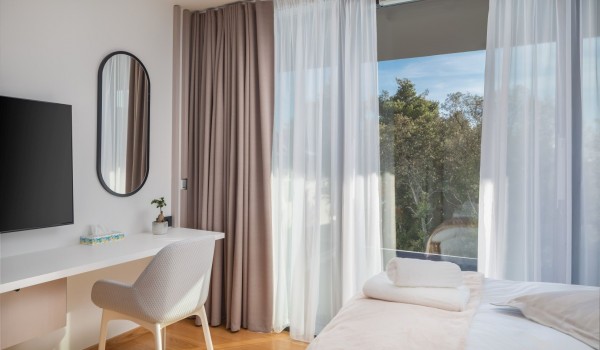 Air conditioned bedroom with king size bed, LCD TV, a table with a chair and oval mirror on the wall and elegant beige toned curtains covering glass wall