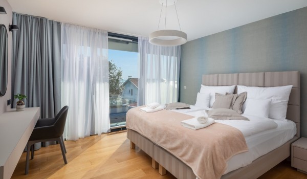An elegant bedroom with en suite bathroom, a table with a chair and oval mirror, and floor-to-ceiling glass wall with a view on sea and greenery surrounding Luxury Villa Idria Sea and View Pe