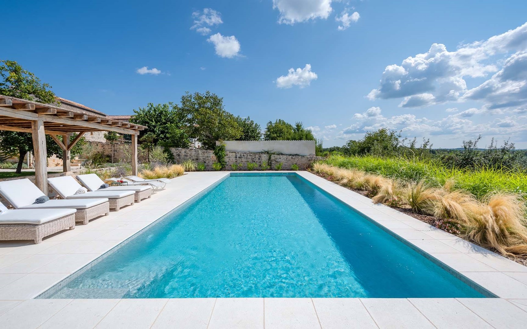 Blick auf den von Sonnenliegen umgebenen Pool auf einer gepflasterten Terrasse im privaten Garten voller Grün vor der Luxusvilla Deluxe Manor Baderna in Istrien