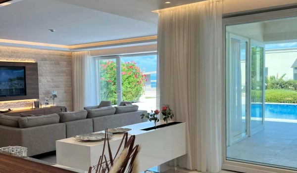 View from the dining area to the comfortable living room with a large corner sofa and two armchairs and a large glass wall that separates the interior from the sunny terrace with the pool