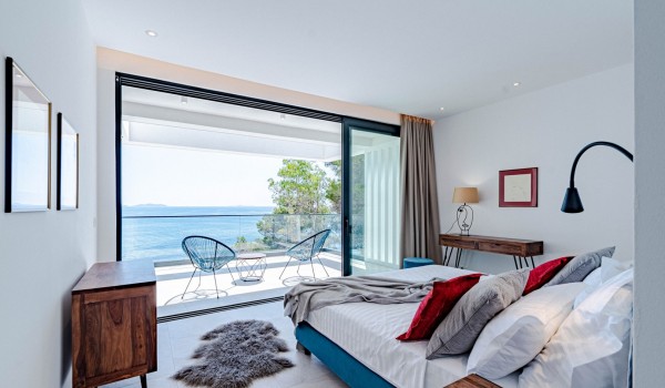 Luxury Master bedroom with a king-size bed, floor-to-ceiling glass walls and a magnificent balcony with a panoramic sea view, coffee table, and two chairs