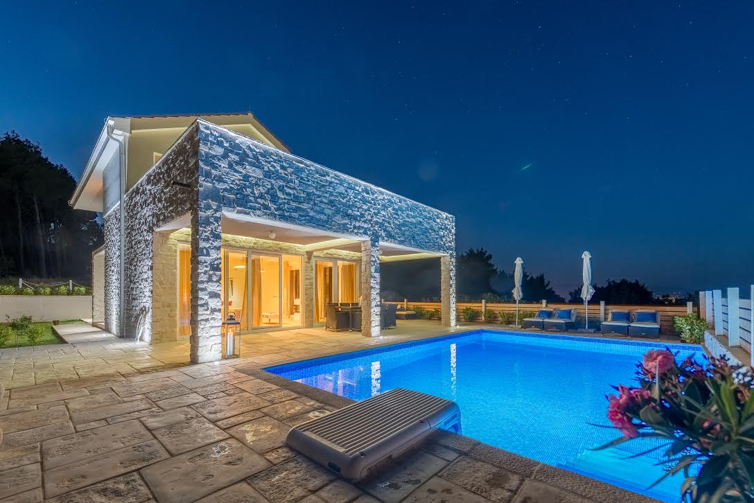 Paved terrace with a private illuminated pool and an outdoor dining table with chairs in the roofed part of the terrace in front of the entrance to the luxury Croatian villa for rent Hvar Lav