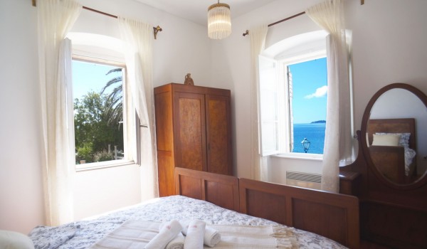 A bedroom with a double bed, a wooden dresser with a mirror, a wooden wardrobe and a window with a view of the sea