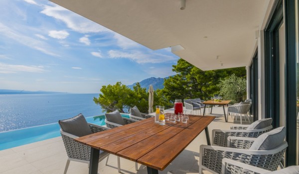 Outdoor covered terrace with a wooden table, chairs and a view of the private heated pool and the sea in a luxury villa for rent in Brela