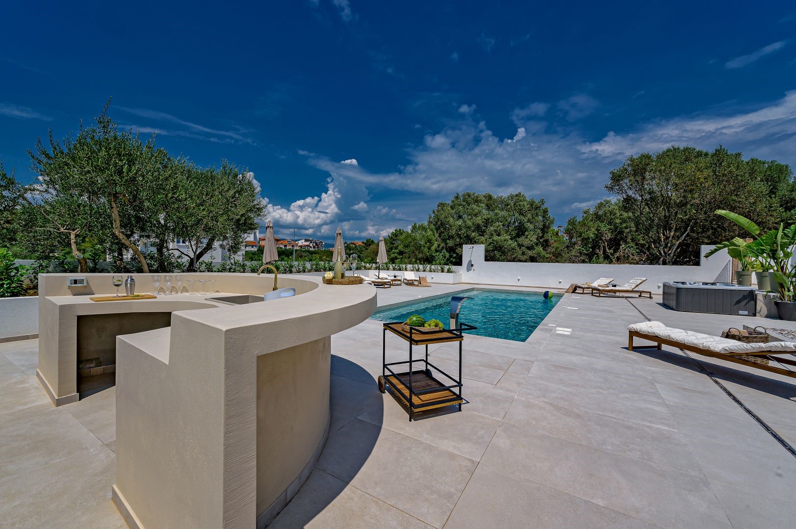 View of the summer kitchen and swimming pool surrounded by comfortable sunbeds and umbrellas on the property of the Croatian luxury villa for rent